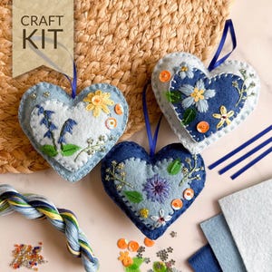 May include: A craft kit featuring three heart-shaped felt ornaments with floral embroidery. The ornaments are in shades of blue and white, with yellow and orange accents. The kit includes embroidery floss, sequins, and felt fabric. The text "CRAFT KIT" is visible.