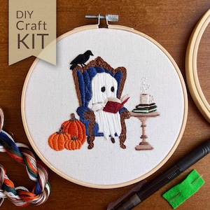Completed Bookworm Ghost hand embroidery filled in with soft white, black and warm autumn tones— a cute ghost reading a book in a cozy dark blue armchair with a raven perched on the back. Next to the chair is a decorate table with a stack of books and steaming cup of hot chocolate. On the other side are three orange pumpkins. The design makes you feel all warm and cozy, perfect decor for spooky season!