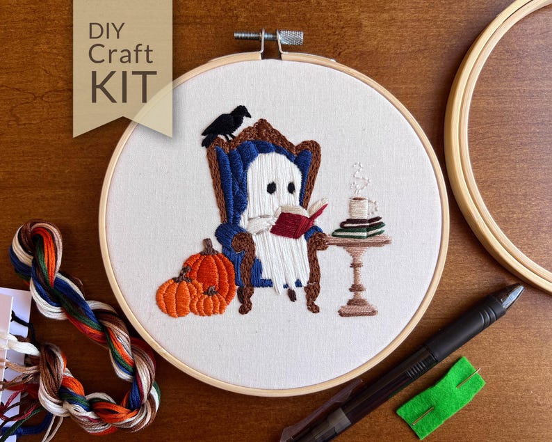 Completed Bookworm Ghost hand embroidery filled in with soft white, black and warm autumn tones— a cute ghost reading a book in a cozy dark blue armchair with a raven perched on the back. Next to the chair is a decorate table with a stack of books and steaming cup of hot chocolate. On the other side are three orange pumpkins. The design makes you feel all warm and cozy, perfect decor for spooky season!