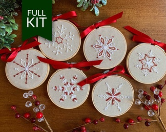 May include: Six unfinished embroidery hoops with red ribbon and white and red snowflake designs. The hoops are on a wooden surface with red berries and greenery.