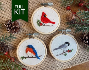 May include: Three embroidery hoop ornaments with winter bird designs. The hoops are made of wood and have a white fabric background. The birds are a cardinal, a bluebird, and a chickadee. Each bird is embroidered with colorful thread and sits on a branch with berries.