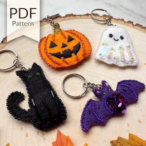 May include: A collection of Halloween-themed keychains. Includes a black cat, a jack-o'-lantern, a ghost, and a purple bat, all made of felt and embellished with beads. The keychains are attached to silver key rings.