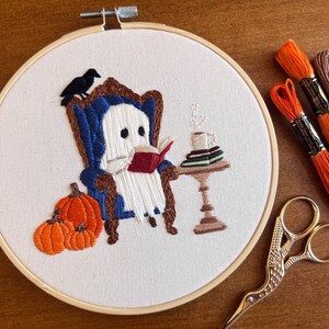 Embroidered hoop art with a white ghost sitting in a blue armchair reading a red book. There are three orange pumpkins in front of the ghost. The ghost is sitting in front of a small table with a cup of coffee and a stack of books. The embroidery is on a white background with a wooden hoop.