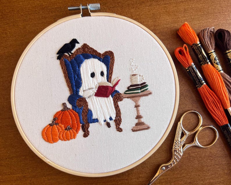 Embroidered hoop art with a white ghost sitting in a blue armchair reading a red book. There are three orange pumpkins in front of the ghost. The ghost is sitting in front of a small table with a cup of coffee and a stack of books. The embroidery is on a white background with a wooden hoop.