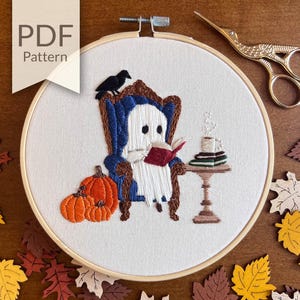 May include: Embroidered hoop art featuring a ghost sitting in a blue armchair with a book and a cup of coffee on a table. The ghost is wearing a white sheet and has a friendly face. There are fall leaves scattered around the hoop.