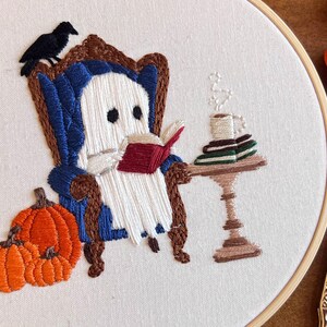Close up detail of a finished embroidered hoop art with a white ghost sitting in a blue armchair reading a red book. The ghost is holding a cup of coffee and there are books stacked on a small table next to the chair. There are two orange pumpkins in the bottom left corner of the hoop. This image shows the finished project that you will get all of the craft supplies to make.