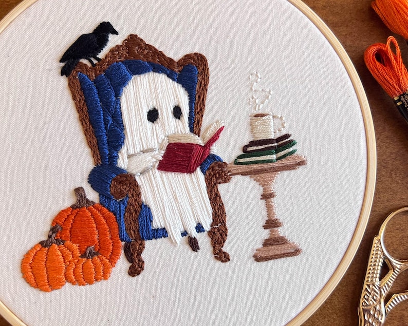 Close up detail of a finished embroidered hoop art with a white ghost sitting in a blue armchair reading a red book. The ghost is holding a cup of coffee and there are books stacked on a small table next to the chair. There are two orange pumpkins in the bottom left corner of the hoop. This image shows the finished project that you will get all of the craft supplies to make.