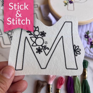 May include: A white felt letter M with a black outline and floral embroidery. The letter is held in a hand and surrounded by embroidery floss in various colors. The text "Stick & Stitch" is visible in the background.