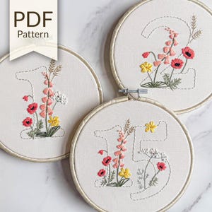 May include: Three embroidery hoops with floral designs. Each hoop features a number: 1, 2, and 5, embroidered with colorful flowers in shades of red, pink, and yellow. The text "PDF Pattern" is visible on the top left.