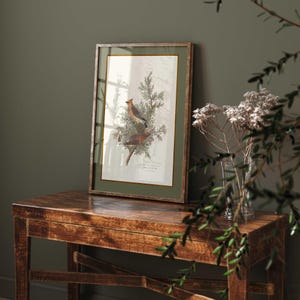 May include: A framed botanical print depicting two birds on a branch, with a green border and a wooden frame. The print is displayed on a rustic wooden table. A vase of dried flowers and greenery are in the foreground.