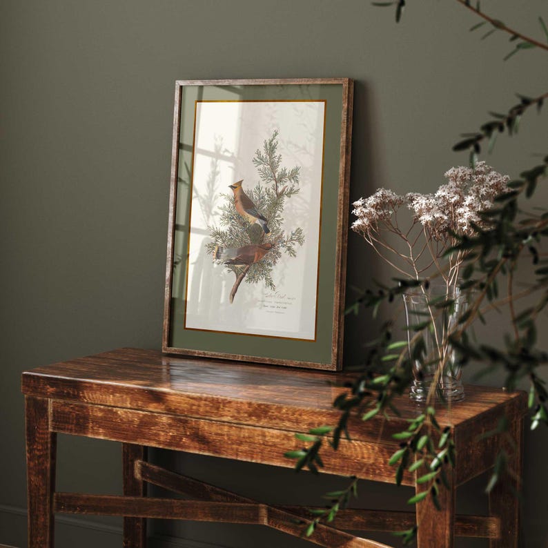 May include: A framed botanical print depicting two birds on a branch, with a green border and a wooden frame. The print is displayed on a rustic wooden table. A vase of dried flowers and greenery are in the foreground.