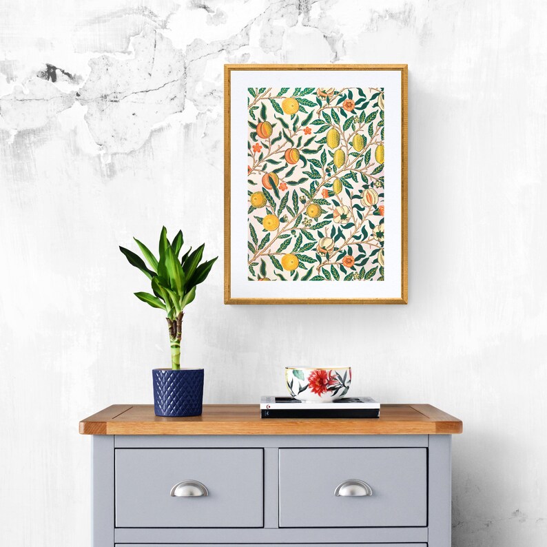 May include: A framed print of a vintage floral design featuring a repeating pattern of green leaves, yellow lemons, and orange flowers. The print is displayed on a light grey wall above a wooden dresser with a blue ceramic planter and a plant.