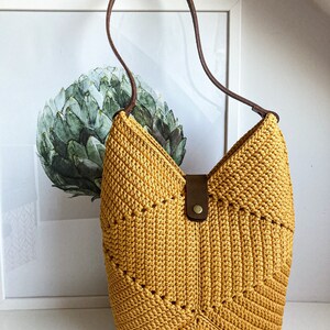 Op de afbeelding: Een gele gehaakte tote bag met een bruin leren band en een bruin leren gesp. De tas heeft een geometrisch patroon.