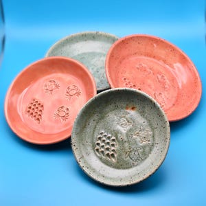 May include: Four handmade ceramic plates in shades of coral, seafoam green, and speckled gray. Each plate features carved designs of crabs, turtles, or honeycomb patterns. The plates are arranged on a bright blue surface.