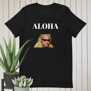 Pode incluir: Camiseta preta com a palavra "ALOHA" impressa em branco na frente. Uma foto de um homem com cabelo loiro e óculos de sol está impressa abaixo da palavra.
