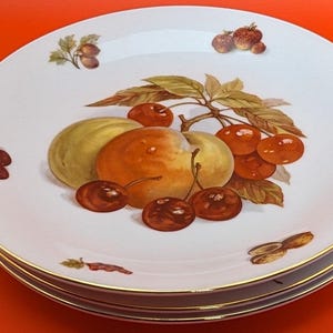 May include: A set of three white ceramic plates with a gold rim. The plates feature a hand-painted design of fruit, including apples, cherries, and plums.