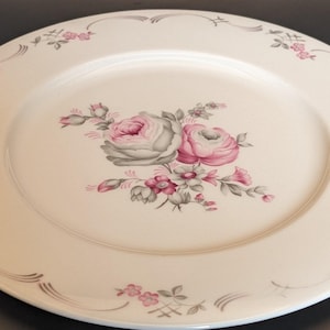 May include: A white ceramic dinner plate with a gray and pink floral design. The plate has a scalloped edge and a delicate floral pattern around the rim.