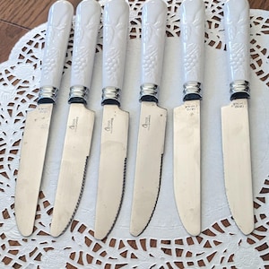 May include: Set of six white handled steak knives with serrated blades. The handles have a grapevine design and the blades are stamped with "Alison Homeware" and "Made in China".