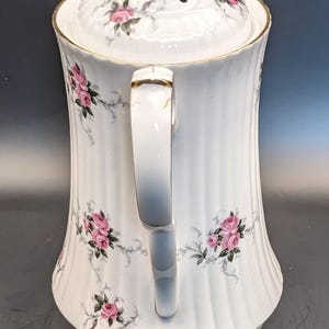 May include: White ceramic teapot with a gold trim and a floral design featuring pink roses. The teapot has a lid with a small hole in the top and a handle on the side.