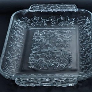 May include: A clear glass baking dish with a floral design. The dish has two handles on the sides and a raised rim.