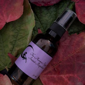 May include: A dark brown glass spray bottle of insect repellent with a black spray nozzle and a purple label. The label reads "The Aroma Fairy Insect Repellent" and features a fairy silhouette. The bottle is on red and green leaves.