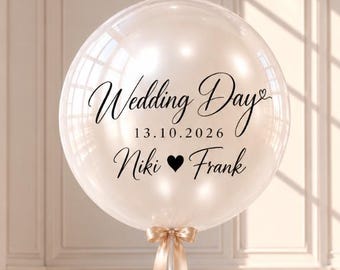 May include: A large, round, translucent cream-colored balloon featuring black text that reads Wedding Day, the date 13.10.2026, and the names Niki and Frank separated by a heart symbol. A light beige ribbon is tied at the base of the balloon.