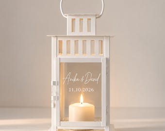 May include: A white metal lantern with a top handle and glass panels. The front glass features the white text Anika & David and the date 11.10.2026. Inside the lantern, a single lit white pillar candle provides a warm glow.