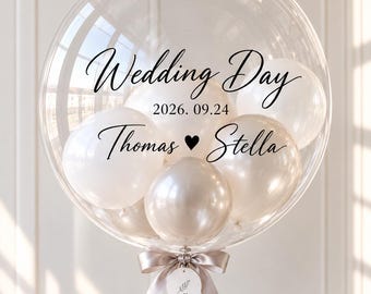 May include: A large clear bubble balloon containing smaller white and cream balloons. The exterior is printed with the black text Wedding Day, the date 2026. 09.24, and the names Thomas and Stella separated by a heart. A satin ribbon bow and a small tag are attached.
