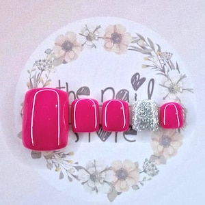 May include: A set of fuchsia-coloured press-on toenails, each with a white curved line design. One nail is covered in silver glitter. The nails are displayed on a floral-themed background.