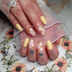 May include: Close-up of a hand with yellow and white nail polish. The nails are decorated with small white flowers and yellow bees.