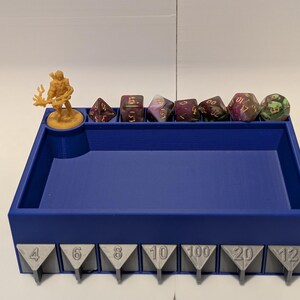 May include: Blue plastic dice tray with compartments for different dice sizes. The tray has a miniature figure in the top left corner and numbers 4, 6, 8, 10, 100, 20, and 12 printed on the front.