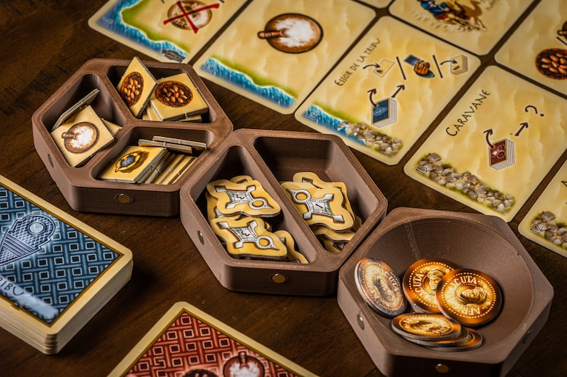 K&ouml;nnte beinhalten: Ein h&ouml;lzerner Spielorganisator mit F&auml;chern zur Aufbewahrung von Spielsteinen. Der Organisator hat eine sechseckige Form und ist aus dunkelbraunem Holz gefertigt. Die F&auml;cher sind mit verschiedenen Spielsteinen gef&uuml;llt, darunter kleine Holzmarker, Karten und M&uuml;nzen. Der Organisator steht auf einem Holztisch.