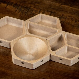 K&ouml;nnte beinhalten: Ein Set aus f&uuml;nf sechseckigen Holzschalen mit hellbrauner Oberfl&auml;che. Die Schalen sind in einem Wabenmuster angeordnet und haben eine glatte, abgerundete Oberfl&auml;che. Jede Schale hat einen kleinen, silberfarbenen Magneten auf der Unterseite.