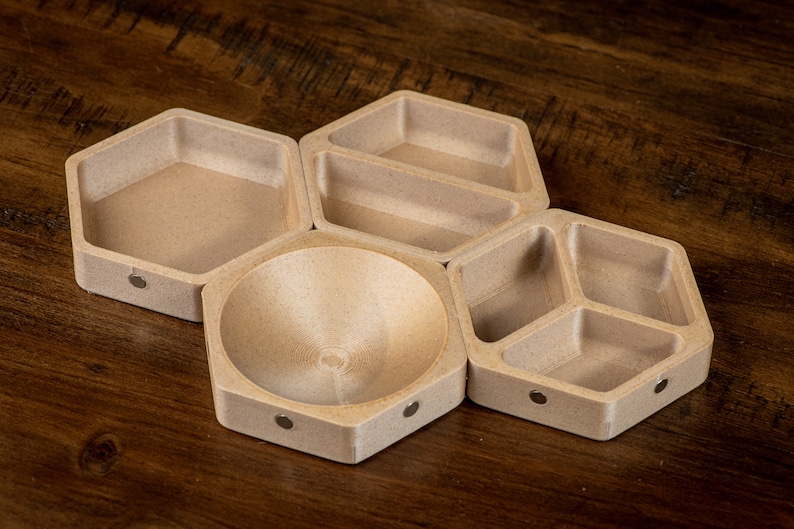 K&ouml;nnte beinhalten: Ein Set aus f&uuml;nf sechseckigen Holzschalen mit hellbrauner Oberfl&auml;che. Die Schalen sind in einem Wabenmuster angeordnet und haben eine glatte, abgerundete Oberfl&auml;che. Jede Schale hat einen kleinen, silberfarbenen Magneten auf der Unterseite.