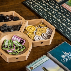 K&ouml;nnte beinhalten: Ein sechseckiges Tablett aus Holz mit drei F&auml;chern. Die F&auml;cher enthalten schwarze und braune Spielsteine, goldene M&uuml;nzen und gr&uuml;ne und rosa Spielkarten mit dem Text "10 PRET" und "30 PRET".