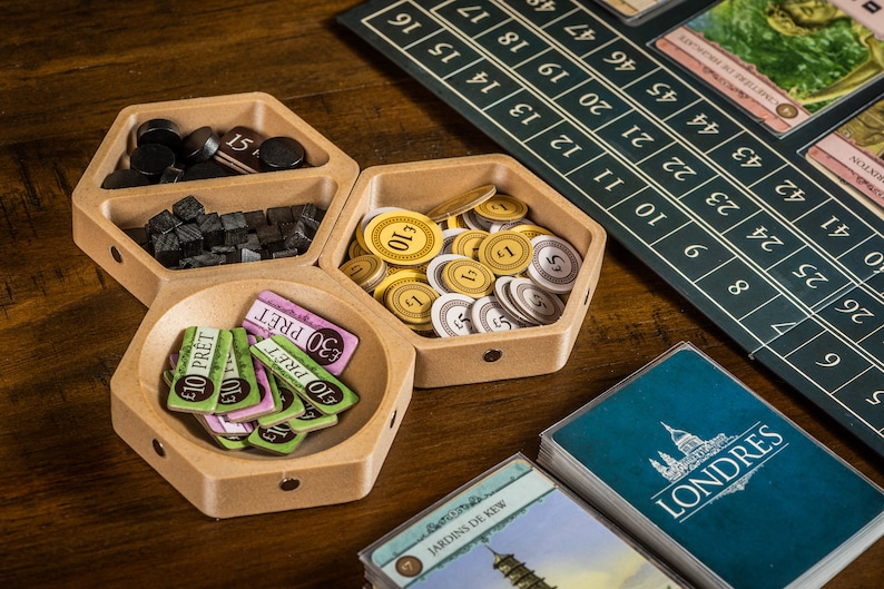 K&ouml;nnte beinhalten: Ein sechseckiges Tablett aus Holz mit drei F&auml;chern. Die F&auml;cher enthalten schwarze und braune Spielsteine, goldene M&uuml;nzen und gr&uuml;ne und rosa Spielkarten mit dem Text "10 PRET" und "30 PRET".