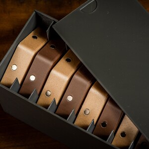 K&ouml;nnte beinhalten: Eine schwarze Box mit offenem Deckel, die einen Stapel brauner und beiger Lederriemen mit silbernen Metallakzenten enth&uuml;llt.