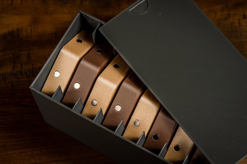 K&ouml;nnte beinhalten: Eine schwarze Box mit offenem Deckel, die einen Stapel brauner und beiger Lederriemen mit silbernen Metallakzenten enth&uuml;llt.