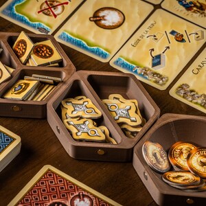 K&ouml;nnte beinhalten: Ein h&ouml;lzerner Spielorganisator mit F&auml;chern zur Aufbewahrung von Spielsteinen. Der Organisator hat eine sechseckige Form und ist aus dunkelbraunem Holz gefertigt. Die F&auml;cher sind mit verschiedenen Spielsteinen gef&uuml;llt, darunter kleine Holzmarker, Karten und M&uuml;nzen. Der Organisator steht auf einem Holztisch.