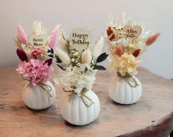 Puede incluir: Tres jarrones blancos con forma de calabaza que contienen arreglos de flores secas. El jarrón de la izquierda tiene flores rosas y moradas con una etiqueta de corazón que dice Beste Mama. El jarrón central tiene flores blancas con una etiqueta redonda que dice Happy Birthday. El jarrón de la derecha tiene flores naranjas y crema con una etiqueta de corazón que dice Alles Gute.