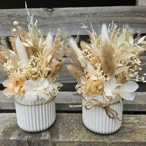 Könnte beinhalten: Zwei weiße Keramikvasen mit gerippter Textur, jede gefüllt mit einem Trockenblumenarrangement. Die Arrangements enthalten getrocknete Gräser, Pampasgras und weiße und beige Blumen.