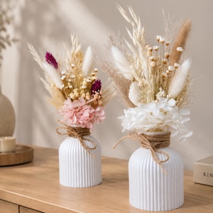 Könnte beinhalten: Zwei weiße, gerippte Keramikvasen, jeweils mit einer braunen Bindfaden-Schleife am Hals. Die linke Vase enthält getrocknete Blumen in Rosa, Magenta und Creme. Die rechte Vase ist mit getrockneten weißen Blumen und Gräsern gefüllt.