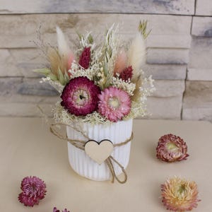 Könnte beinhalten: Eine weiße, gerippte Keramikvase enthält ein Trockenblumenarrangement. Das Arrangement umfasst rosa, bordeauxfarbene und cremefarbene Blumen und Gräser. Ein Holzherz ist mit Bindfaden an der Vase befestigt. Zusätzliche Trockenblumen sind verstreut.