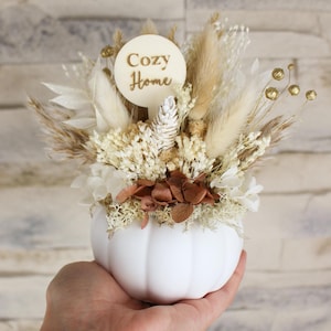 Könnte beinhalten: Eine weiße, kürbisförmige Vase gefüllt mit getrockneten Blumen und Blättern in Creme-, Beige- und Brauntönen. Eine Holzscheibe mit den Worten "Cozy Home" befindet sich in der Anordnung. Die Vase wird in einer Hand gehalten.