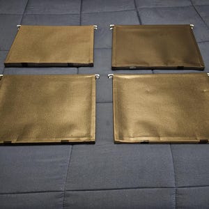 May include: Four brown file organizers with metal hanging tabs are arranged on a blue quilted surface. The organizers are rectangular and appear to be made of a durable, textured material. The tabs are white and are attached to the top edge of each organizer.