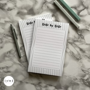 May include: Two white notepad sets with gray stripes and the text "little by little" printed in black. The notepads are on a white marble surface.