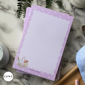 May include: A stack of lavender-colored notepads with a scalloped border and white polka dots. Each notepad features a picnic basket illustration in the bottom corner and the text "YOUR NAME THE LABEL".
