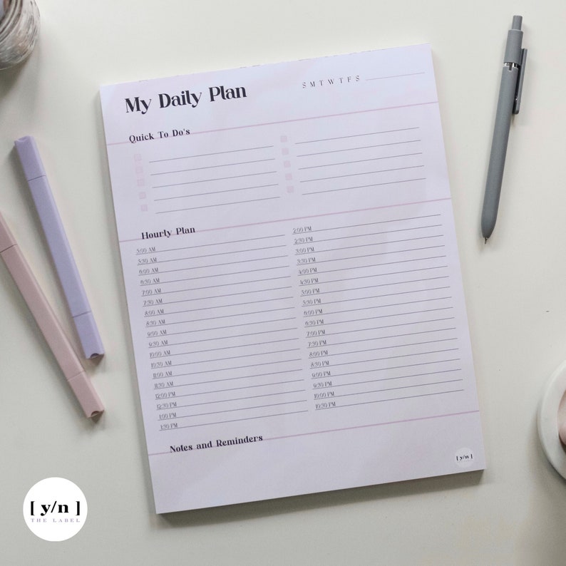 May include: A white daily planner with a pink and gray color scheme. The planner has a section for quick to-dos, an hourly plan, and notes and reminders. The planner has the text "My Daily Plan" at the top.