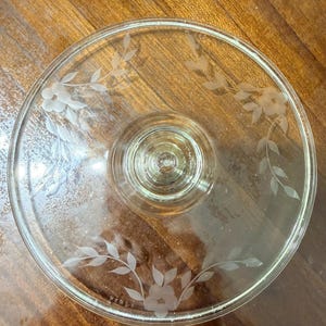 May include: A clear glass cake stand featuring a circular top with a central pedestal base. The glass surface is decorated with three etched floral motifs, each consisting of a five-petaled flower surrounded by delicate leafy vine patterns.