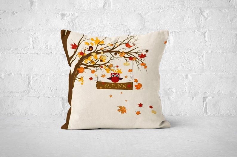 Pu&ograve; includere: Un cuscino decorativo con sfondo bianco che presenta un albero marrone con foglie arancioni e gialle. Un gufo &egrave; appollaiato su un ramo con un cartello che dice "Autunno".